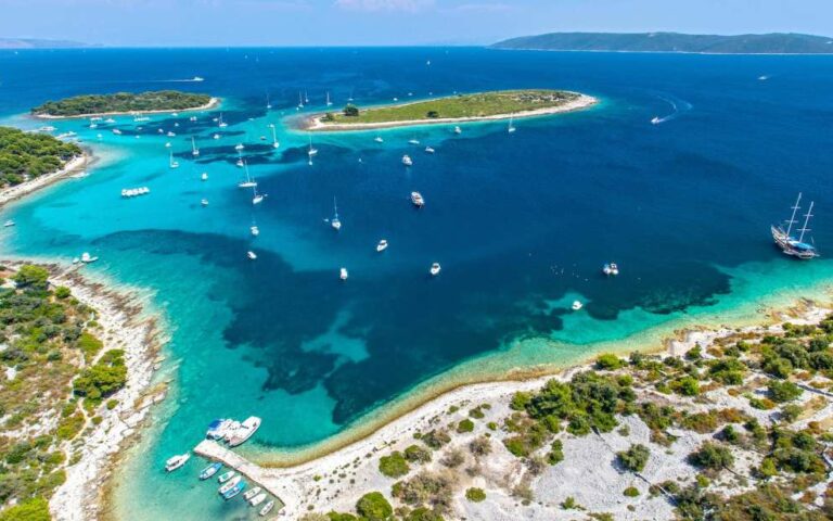 Luftaufnahme einer wunderschönen Küstenregion mit klarem, blauem Wasser, mehreren kleinen Inseln und zahlreichen vor Anker liegenden Booten und Yachten. Grüne Vegetation und felsiges Gelände umgeben die Küste, und auf dem Land sind einige Gebäude zu sehen.