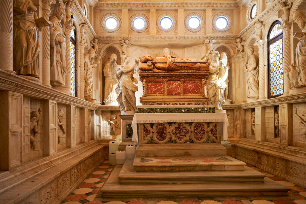 Ein reich verziertes Grabmal in einer Kathedrale mit einem großen Sarkophag aus rotem und weißem Marmor, umgeben von detailreichen Skulpturen von Heiligen und Engeln. Der Raum wird durch Buntglasfenster beleuchtet und von aufwendigen Wandschnitzereien und Säulen eingerahmt.