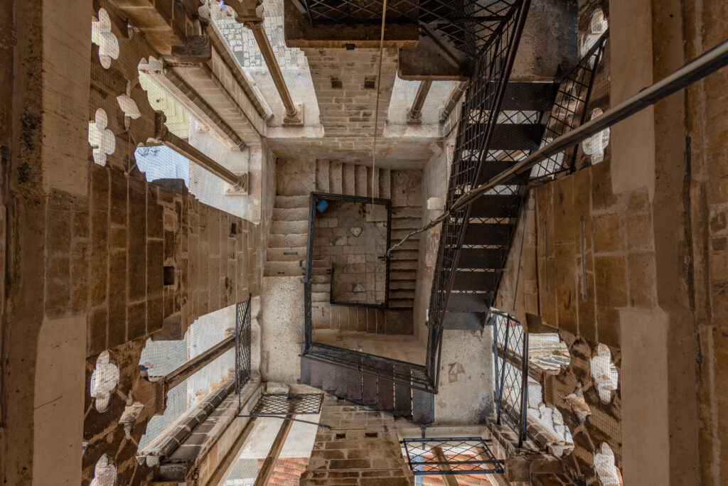 Eine Wendeltreppe führt in einen verwitterten Steinturm hinauf, flankiert von dekorativen Fenstern mit aufwendigen Ausschnitten. Die Treppen und Podeste sind aus Metall und Stein gefertigt und vereinen in diesem architektonischen Interieur rustikale und industrielle Elemente.