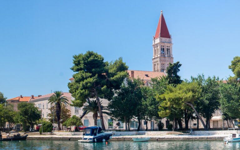 Panoramablick auf eine ruhige Küstenstadt mit dem Turm der Kathedrale St. Laurentius, dessen rotes Dach in den blauen Himmel ragt. Im Vordergrund sind Boote zu sehen, die an einer Uferpromenade angedockt sind, die von üppigen Bäumen und historischen Gebäuden gesäumt ist, und das bei hellem Sonnenlicht.