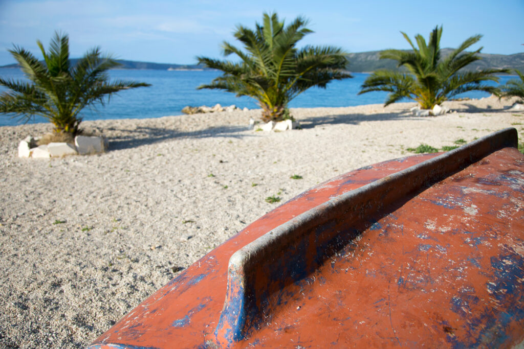 Ein verwittertes orange-blaues Boot liegt kopfüber auf einem Sandstrand mit Kieselsteinen. Vier Palmen im Hintergrund blicken auf ein ruhiges, blaues Meer unter einem klaren Himmel. Die Szene an den Stränden ist ruhig und sonnig und zeigt eine Küstenlandschaft.