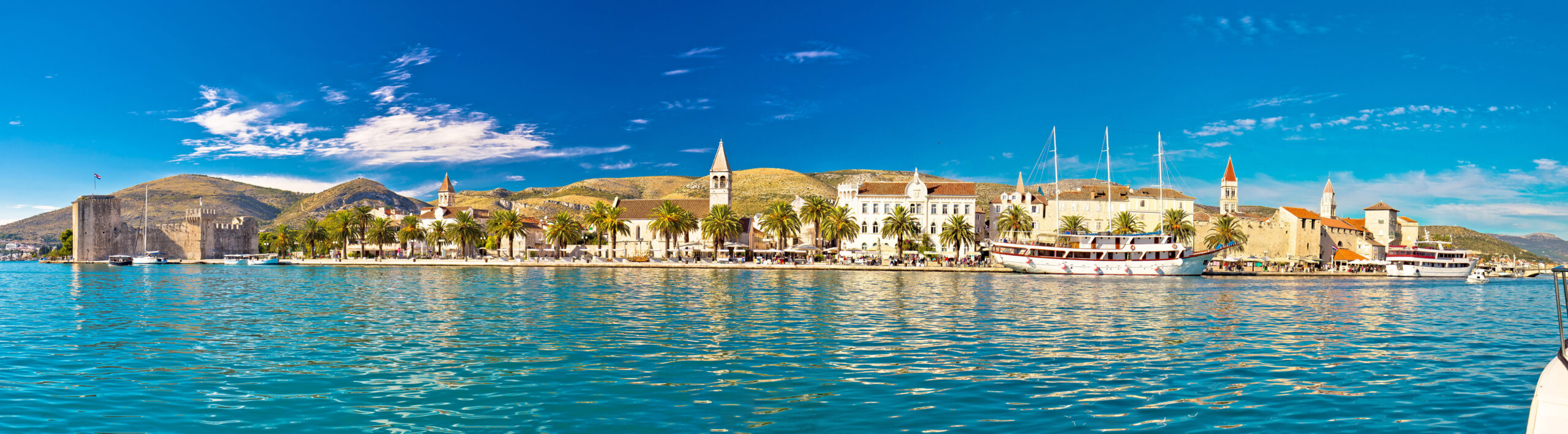 Panoramablick auf eine Küstenstadt am Mittelmeer mit historischen Steingebäuden, Palmen und Kirchtürmen unter einem klaren blauen Himmel. Boote liegen am Ufer festgemacht, im Hintergrund erheben sich Hügel. Das ruhige Meer reflektiert die leuchtenden Farben und macht es zu einem perfekten Urlaubsziel.