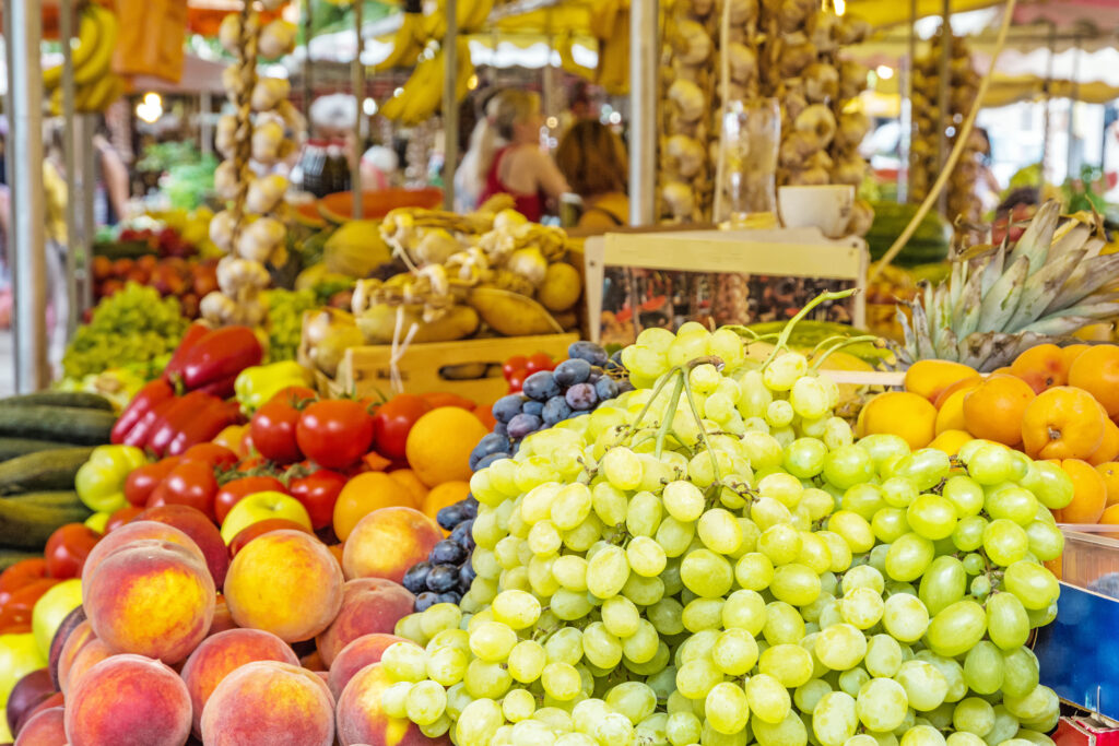 Ein lebhafter Marktstand mit einer Vielzahl frischer Produkte, darunter im Vordergrund grüne und schwarze Weintrauben, Pfirsiche, Tomaten und Orangen. Im Hintergrund sind Knoblauchschnüre und andere Obst- und Gemüsesorten ausgestellt.