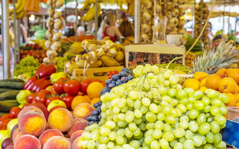 Ein lebhafter Marktstand mit einer Vielzahl frischer Produkte, darunter im Vordergrund grüne und schwarze Weintrauben, Pfirsiche, Tomaten und Orangen. Im Hintergrund sind Knoblauchschnüre und andere Obst- und Gemüsesorten ausgestellt.