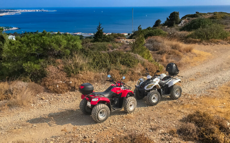 Zwei Geländefahrzeuge (ATVs), ein rotes und ein weißes, stehen auf einem holprigen Feldweg mit Blick auf die Küste. Im Hintergrund ist das Meer mit einem klaren blauen Himmel zu sehen, während der Weg auf beiden Seiten von Sträuchern und Bäumen gesäumt ist.