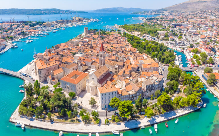Luftaufnahme einer historischen Küstenstadt, die sich perfekt für einen erholsamen Urlaub eignet. Sie besteht aus Gebäuden mit Terrakotta-Dächern auf einer kleinen Halbinsel, umgeben von türkisfarbenem Wasser. An den Ufern liegen Boote vor Anker, und eine Steinbrücke verbindet die Stadt mit dem Festland. Im Hintergrund sind grüne Hügel und andere Siedlungen zu sehen.