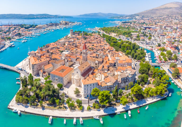 Luftaufnahme einer historischen Küstenstadt, die sich perfekt für einen erholsamen Urlaub eignet. Sie besteht aus Gebäuden mit Terrakotta-Dächern auf einer kleinen Halbinsel, umgeben von türkisfarbenem Wasser. An den Ufern liegen Boote vor Anker, und eine Steinbrücke verbindet die Stadt mit dem Festland. Im Hintergrund sind grüne Hügel und andere Siedlungen zu sehen.