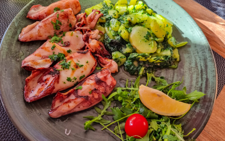 Ein Teller mit gegrillten Calamari, garniert mit gehackten Kräutern, dazu gebratene Kartoffeln mit Gemüse. Dazu gibt es einen kleinen Salat aus Rucola, einer Kirschtomate und einer Zitronenscheibe. Der Teller steht auf einem Holztisch.