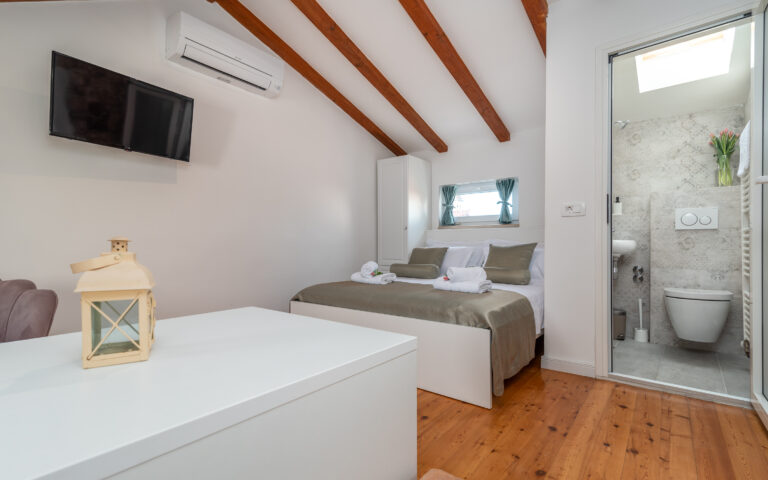 Ein gemütliches Schlafzimmer mit einem Doppelbett mit grün-weißer Bettwäsche, zwei aufgerollten Handtüchern und zwei kleinen Fenstern. Das Zimmer verfügt über Holzbalken an der Decke, einen an der Wand montierten Fernseher, eine Klimaanlage und ein eigenes Badezimmer mit modernen Armaturen. Eine weiße Laterne schmückt einen Tisch im Vordergrund.