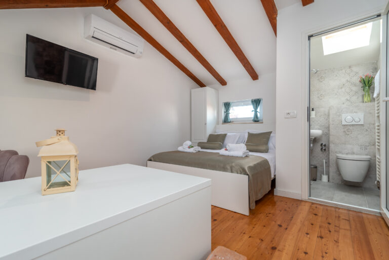 Ein gemütliches Schlafzimmer mit einem Doppelbett mit grün-weißer Bettwäsche, zwei aufgerollten Handtüchern und zwei kleinen Fenstern. Das Zimmer verfügt über Holzbalken an der Decke, einen an der Wand montierten Fernseher, eine Klimaanlage und ein eigenes Badezimmer mit modernen Armaturen. Eine weiße Laterne schmückt einen Tisch im Vordergrund.