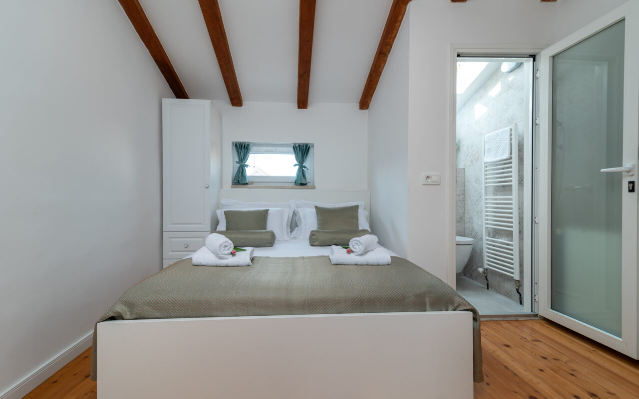 Ein gemütliches Schlafzimmer mit einem Doppelbett, ordentlich gemacht mit weißer Bettwäsche und zwei gefalteten Handtüchern am Fußende. Dekorative Kissen schmücken das Bett. Sichtbare Holzbalken betonen die Decke. Über dem Bett befindet sich ein schmales Fenster mit Vorhängen. Eine angrenzende offene Tür gibt den Blick auf ein Badezimmer frei.