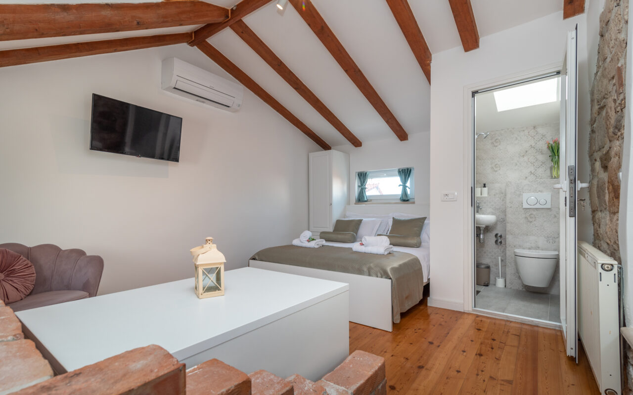 Ein gemütliches Schlafzimmer mit schräger Holzbalkendecke und einem Doppelbett mit beiger und weißer Bettwäsche. Durch eine offene Tür ist ein kleines Badezimmer sichtbar. Das Zimmer verfügt über einen an der Wand montierten Fernseher, eine Klimaanlage und einen kleinen Sitzbereich mit einem rosa gepolsterten Stuhl.