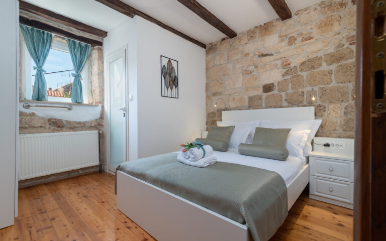 Ein gemütliches Schlafzimmer mit Steinwänden und Holzbalken an der Decke. Das Zimmer ist mit einem Doppelbett mit grauer Bettwäsche, weißen Nachttischen und einem ordentlich gefalteten Handtuch und einer Pflanze auf dem Bett ausgestattet. Ein Fenster mit blauen Vorhängen lässt Tageslicht herein, perfekt für einen erholsamen Urlaub. Eine Tür führt zu einem weiteren Zimmer.