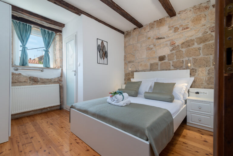 Ein gemütliches Schlafzimmer mit Steinwänden und Holzbalken an der Decke. Das Zimmer ist mit einem Doppelbett mit grauer Bettwäsche, weißen Nachttischen und einem ordentlich gefalteten Handtuch und einer Pflanze auf dem Bett ausgestattet. Ein Fenster mit blauen Vorhängen lässt Tageslicht herein, perfekt für einen erholsamen Urlaub. Eine Tür führt zu einem weiteren Zimmer.