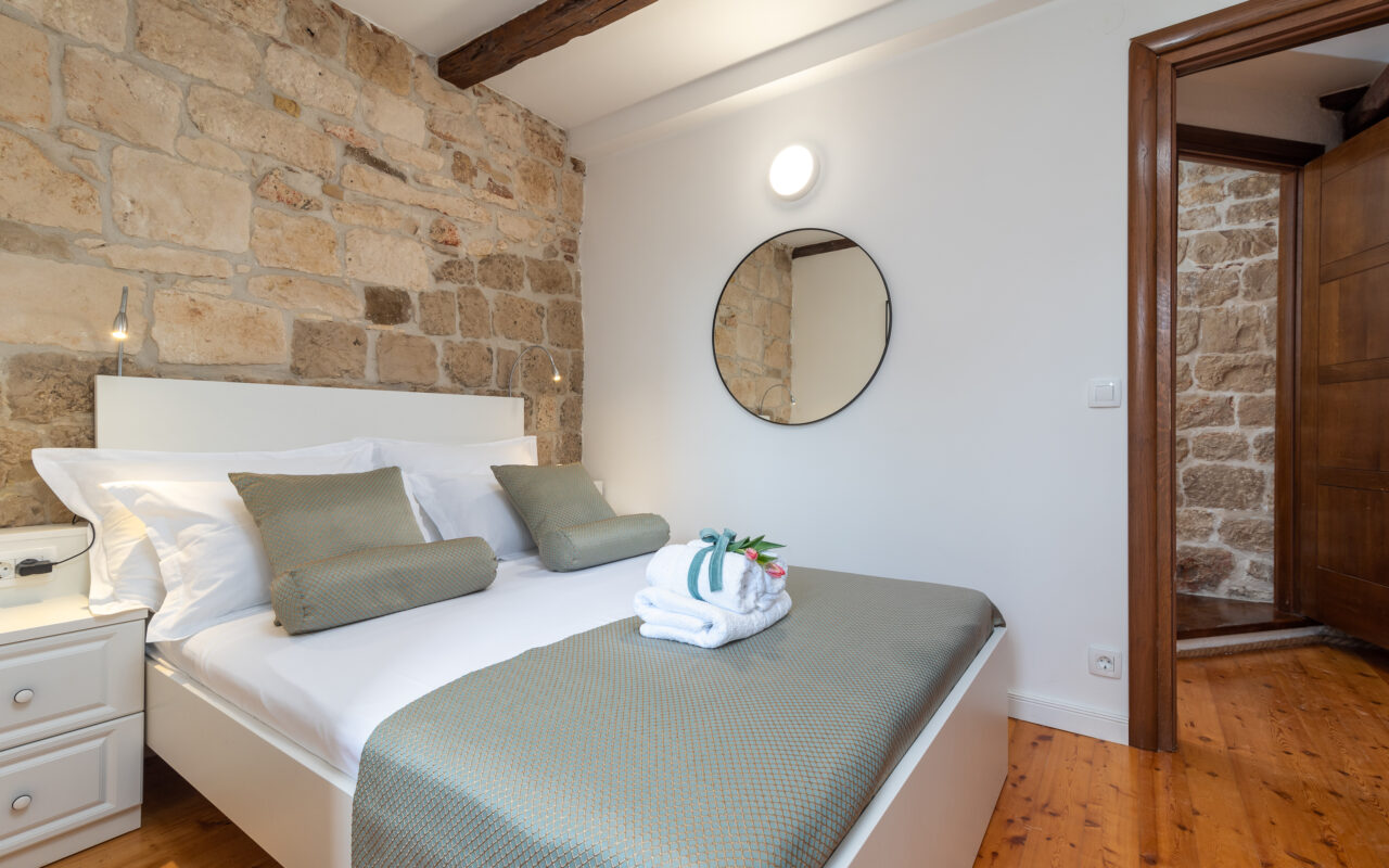 Ein gemütliches Schlafzimmer verfügt über ein großes Bett mit grünen Kissen und einer dazu passenden grünen Decke. Die rustikale Steinwand hinter dem Bett wird durch weiße Wände und hölzerne Deckenbalken kontrastiert. Über dem Bett hängt ein runder Spiegel und auf dem Bett liegen gefaltete Handtücher mit Blumen.