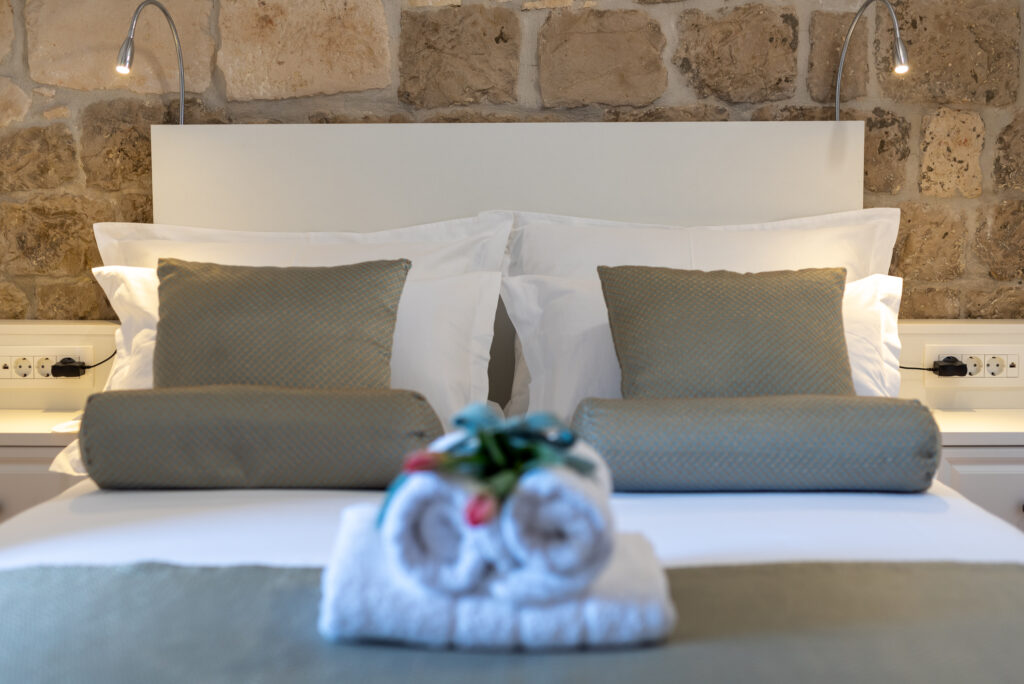 Ein ordentlich gemachtes Bett mit zwei weißen Kissen und zwei zylindrischen grünen Polstern liegt an einer Steinwand. Zwei gerollte weiße Handtücher mit einem dekorativen Blumenmuster liegen am Fußende des Bettes und bieten 5 Gründe, es sich gemütlich zu machen. Am Kopfteil sind verstellbare Leselampen angebracht.