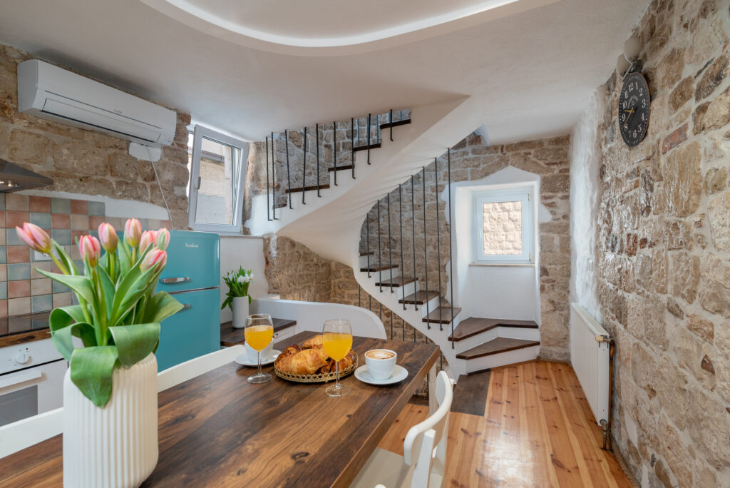 Eine gemütliche Küche mit Steinwänden verfügt über einen hölzernen Esstisch mit Tulpen, Croissants und zwei Gläsern Orangensaft. Die Küche verfügt über einen türkisfarbenen Kühlschrank und eine Treppe, die zu einem Loftbereich führt. Eine große dekorative Uhr schmückt die Wand.