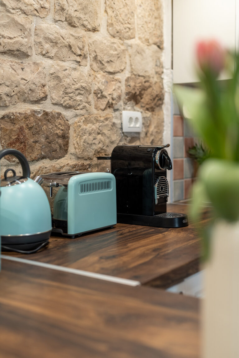 Eine gemütliche Küchentheke mit rustikalem Steinmauerhintergrund, mit einem blaugrünen Wasserkocher, einem dazu passenden blaugrünen Toaster, einer schwarzen Espressomaschine und einem Teil eines Tulpenstraußes im Vordergrund. Die Holzarbeitsplatte verleiht dem einladenden Küchenraum Wärme.