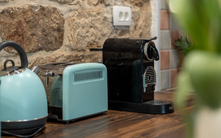 Eine gemütliche Küchentheke mit rustikalem Steinmauerhintergrund, mit einem blaugrünen Wasserkocher, einem dazu passenden blaugrünen Toaster, einer schwarzen Espressomaschine und einem Teil eines Tulpenstraußes im Vordergrund. Die Holzarbeitsplatte verleiht dem einladenden Küchenraum Wärme.