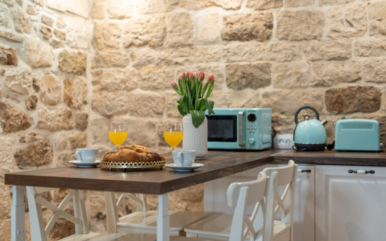 Eine gemütliche Küche mit Backsteinwand verfügt über einen Holztisch mit Croissants, Gläsern mit Orangensaft und Kaffeetassen. Auf der Theke stehen pastellblaue Küchengeräte, darunter ein Toaster, eine Mikrowelle und ein Wasserkocher. Eine Vase mit rosa Tulpen sorgt für einen Hauch von Farbe.