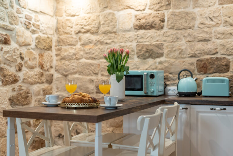 Eine gemütliche Küche mit Backsteinwand verfügt über einen Holztisch mit Croissants, Gläsern mit Orangensaft und Kaffeetassen. Auf der Theke stehen pastellblaue Küchengeräte, darunter ein Toaster, eine Mikrowelle und ein Wasserkocher. Eine Vase mit rosa Tulpen sorgt für einen Hauch von Farbe.