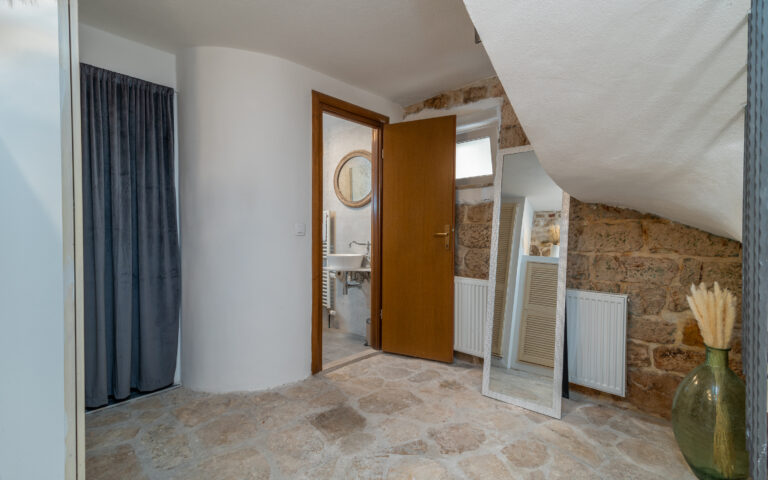 Ein gemütlicher Raum mit geschwungener weißer Wand und Steinboden. Eine offene Holztür gibt den Blick auf ein kleines Badezimmer mit rundem Spiegel frei. Ein Ganzkörperspiegel lehnt an einer abgewinkelten Wand. Eine große Vase mit getrockneten Pflanzen verleiht dem Raum eine dekorative Note.