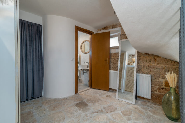 Ein gemütlicher Raum mit geschwungener weißer Wand und Steinboden. Eine offene Holztür gibt den Blick auf ein kleines Badezimmer mit rundem Spiegel frei. Ein Ganzkörperspiegel lehnt an einer abgewinkelten Wand. Eine große Vase mit getrockneten Pflanzen verleiht dem Raum eine dekorative Note.