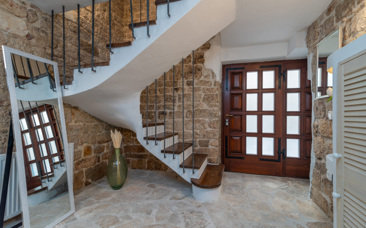 Ein rustikaler Innenraum verfügt über eine Wendeltreppe mit Holzstufen und schwarzem Eisengeländer. Die Wände sind aus Stein und an einer Wand lehnt ein großer Standspiegel. Im Hintergrund ist eine Holztür mit Milchglasplatten zu sehen und neben der Treppe steht eine dekorative Vase.