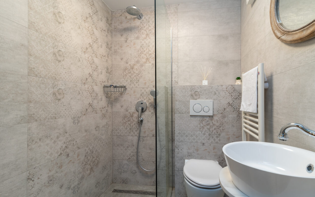 Ein modernes Badezimmer mit ebenerdiger Dusche, abgetrennt durch eine Glaswand, mit wandmontiertem Duschkopf, Chromarmaturen und eingebautem Regal. Daneben befindet sich eine Toilette mit Spülknopf an der Wand, einem runden Waschbecken und einem Spiegel darüber. Der Raum hat geflieste Wände.