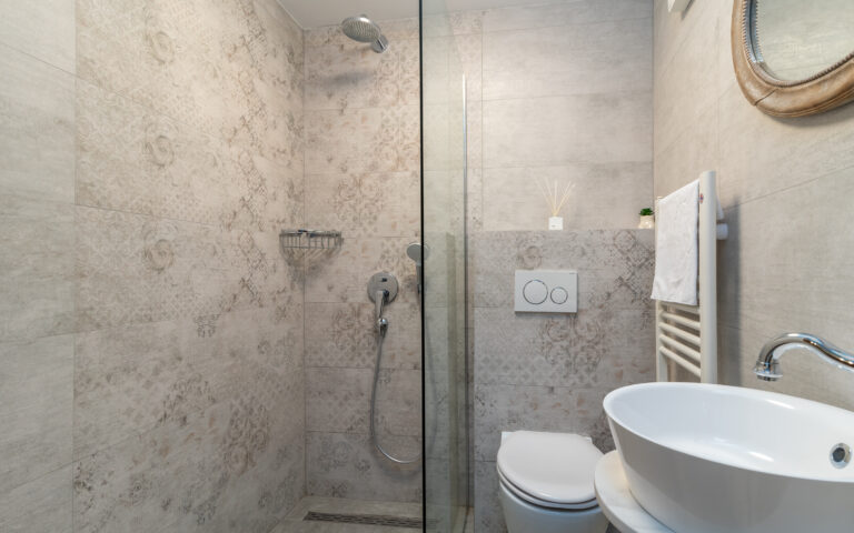 Ein modernes Badezimmer mit ebenerdiger Dusche, abgetrennt durch eine Glaswand, mit wandmontiertem Duschkopf, Chromarmaturen und eingebautem Regal. Daneben befindet sich eine Toilette mit Spülknopf an der Wand, einem runden Waschbecken und einem Spiegel darüber. Der Raum hat geflieste Wände.