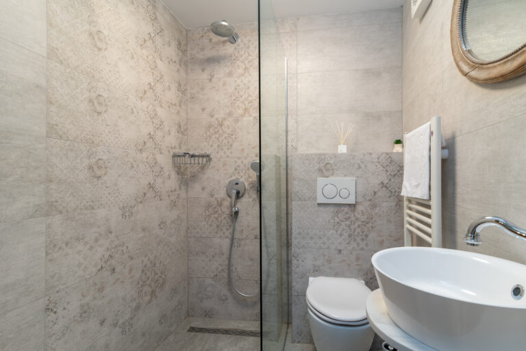 Ein modernes Badezimmer mit ebenerdiger Dusche, abgetrennt durch eine Glaswand, mit wandmontiertem Duschkopf, Chromarmaturen und eingebautem Regal. Daneben befindet sich eine Toilette mit Spülknopf an der Wand, einem runden Waschbecken und einem Spiegel darüber. Der Raum hat geflieste Wände.