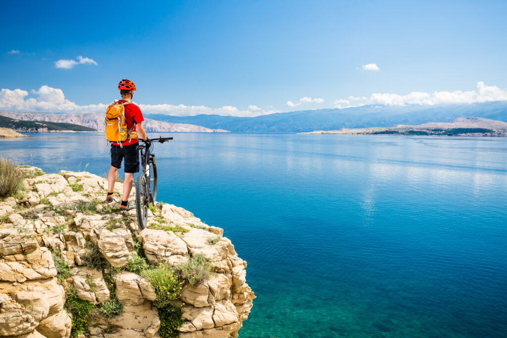 Ein Radfahrer in einem roten Hemd, einem orangefarbenen Helm und einem orangefarbenen Rucksack steht neben einem Mountainbike auf einer Felsklippe und blickt auf einen ruhigen blauen See mit Bergen in der Ferne unter einem klaren Himmel.
