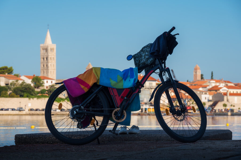 An einem ruhigen Ufer steht ein Fahrrad, das mit einem bunten Handtuch bedeckt ist. Im Hintergrund ist unter einem klaren blauen Himmel eine malerische Stadt mit Gebäuden mit roten Dächern und einem hohen Glockenturm zu sehen.